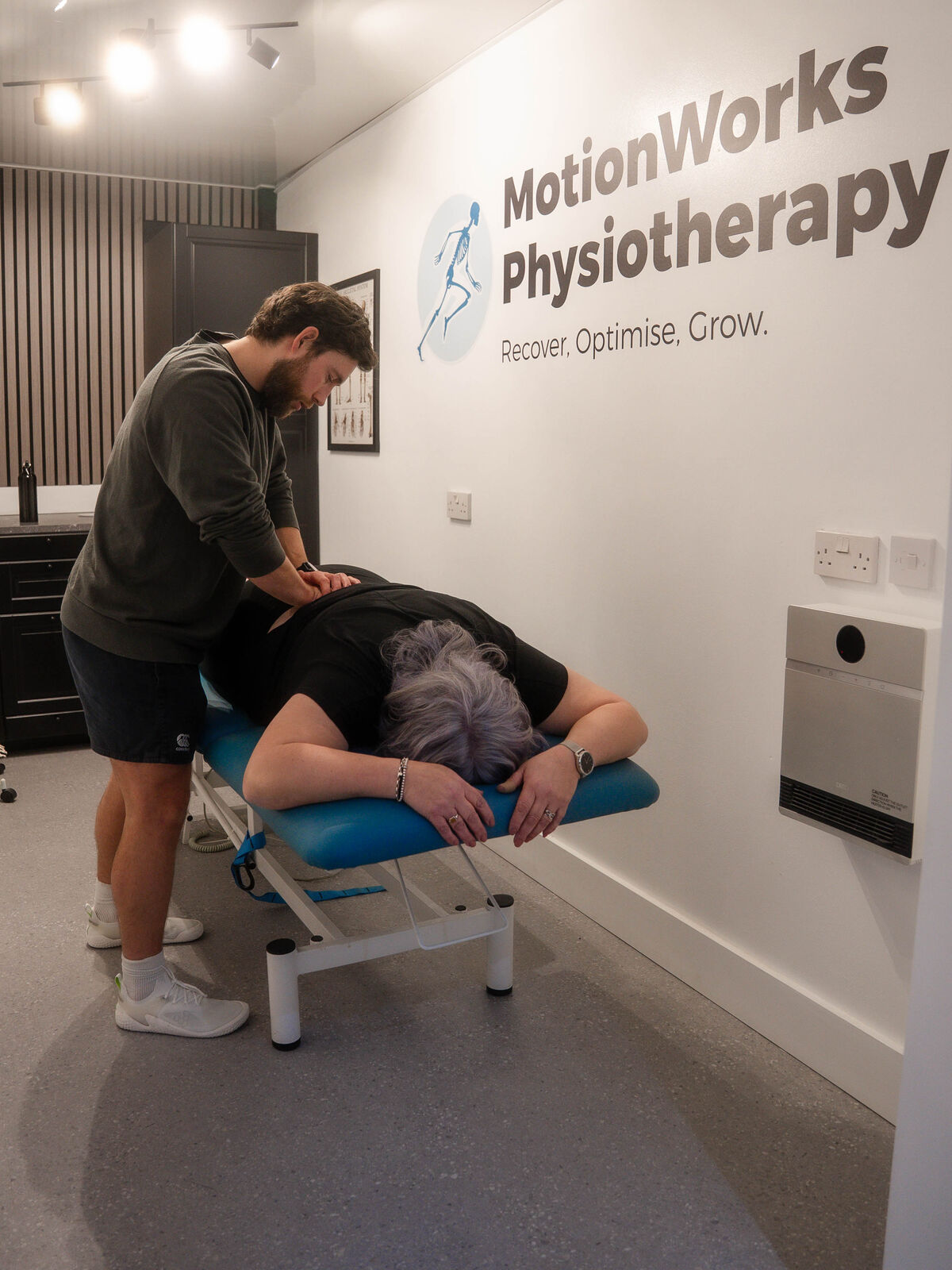 A MotionWorks physiotherapist treating a patient lying on a treatment plinth, with the clinic's wall branding behind them.